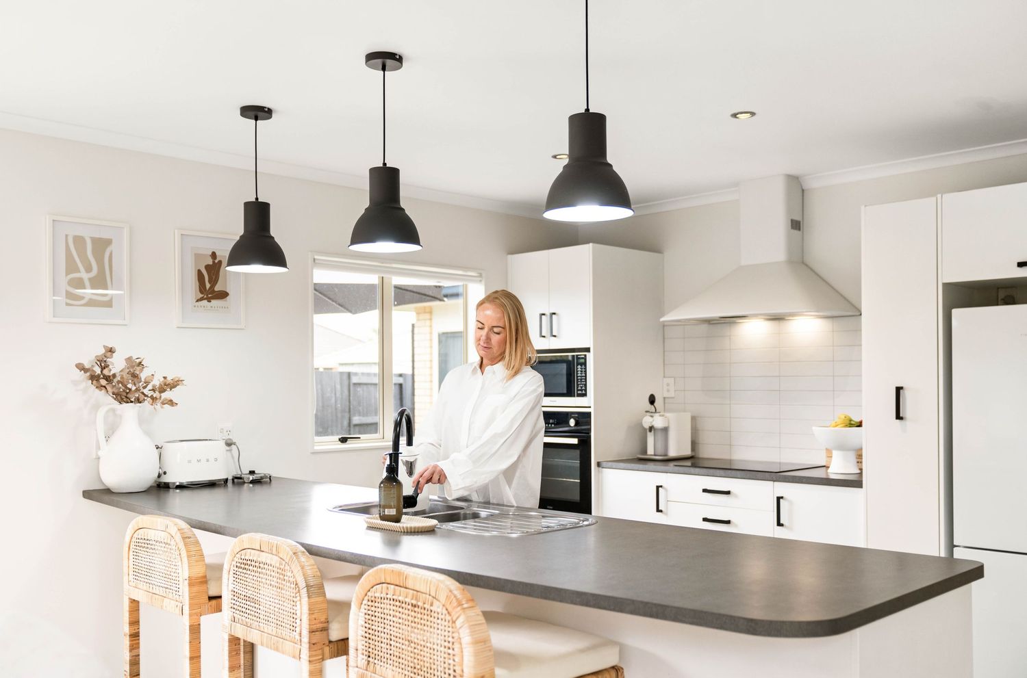 Bright modern kitchen with black pendant lights, rattan bar stools and grey stone countertops in open plan layout.