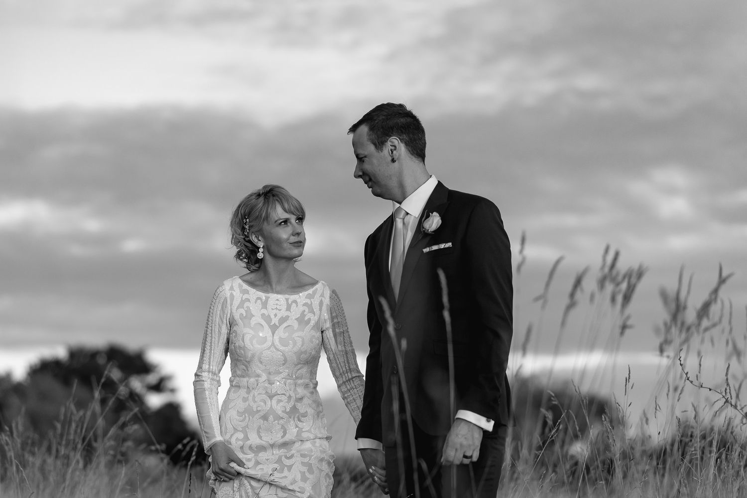 A couple in wedding attire walks together through a scenic field of tall grass during a cloudy afternoon.