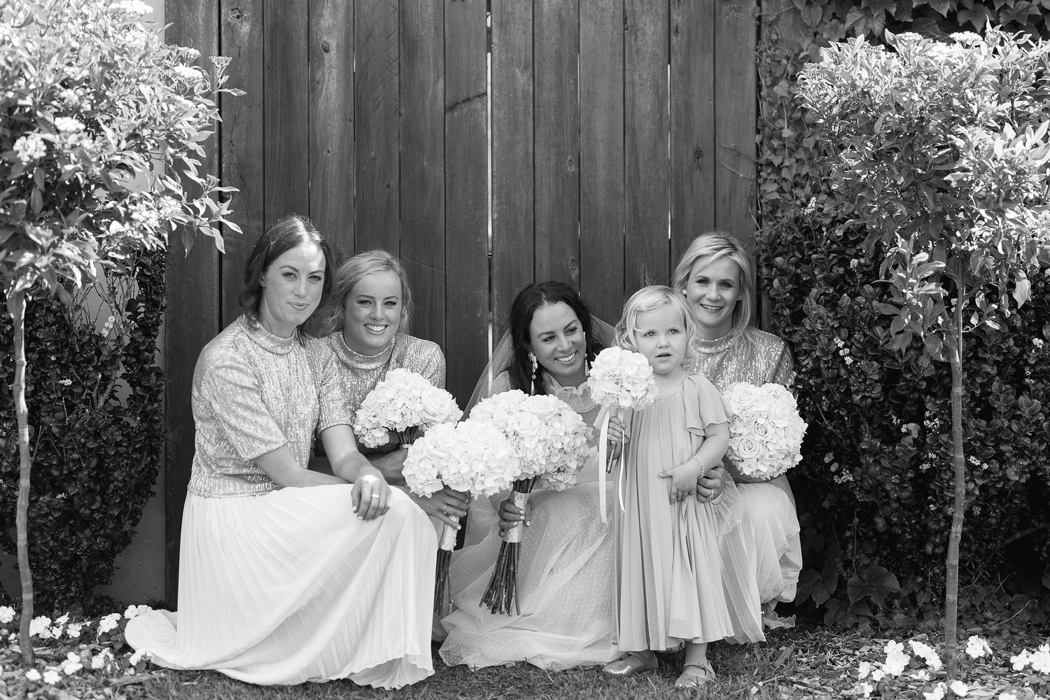Group of people wearing coordinated white outfits pose together in a backyard garden setting with wooden fence.