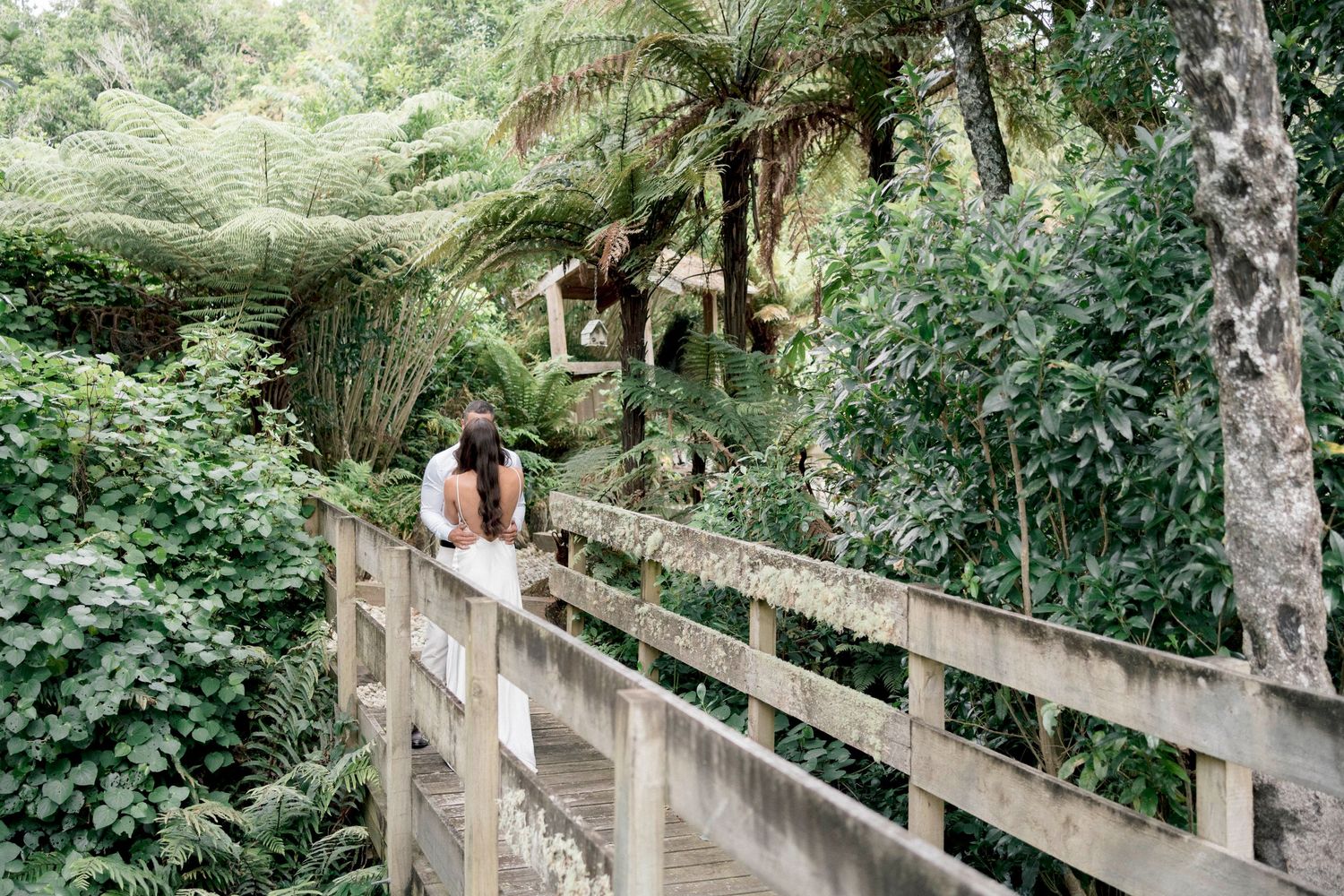 A couple stands on a rustic wooden bridge surrounded by lush tropical plants and palm trees in a peaceful forest setting.
