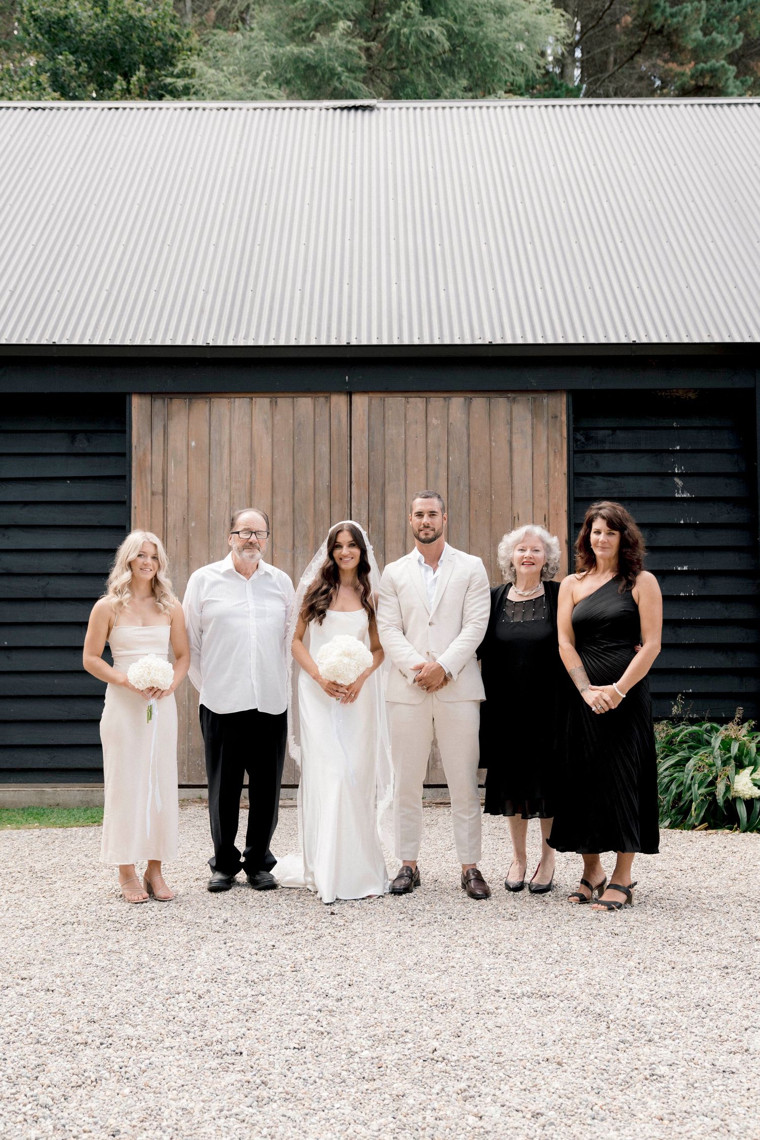 A small group poses together against a black barn with wooden doors during a rustic outdoor wedding ceremony.
