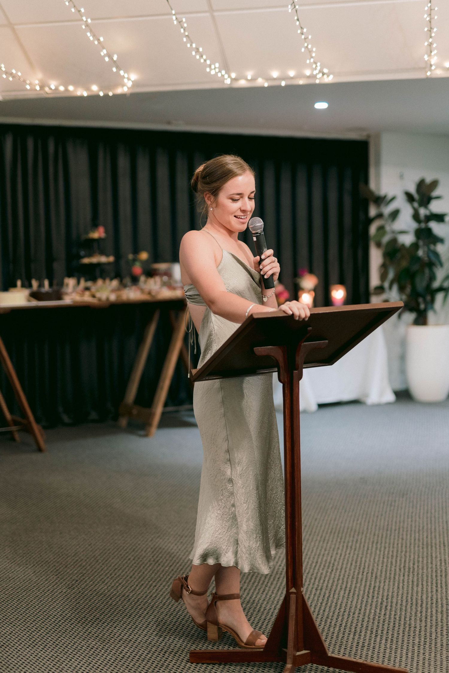 A wedding guest delivers a speech at a reception podium with twinkle lights and dark curtains in the background.