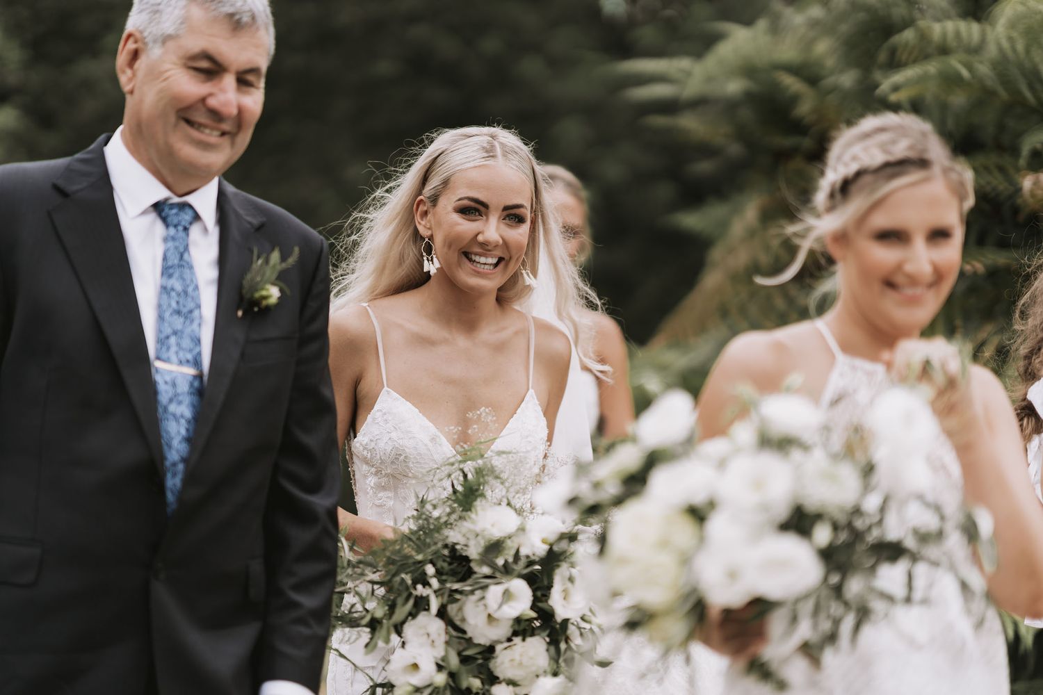 A wedding party walks and smiles together carrying elegant white floral bouquets in an outdoor garden setting.