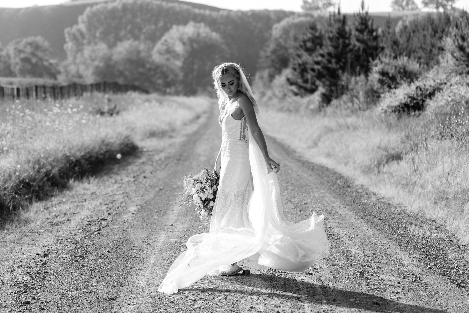A bride in a white wedding dress stands on a rustic country road surrounded by natural scenery and sunlight.