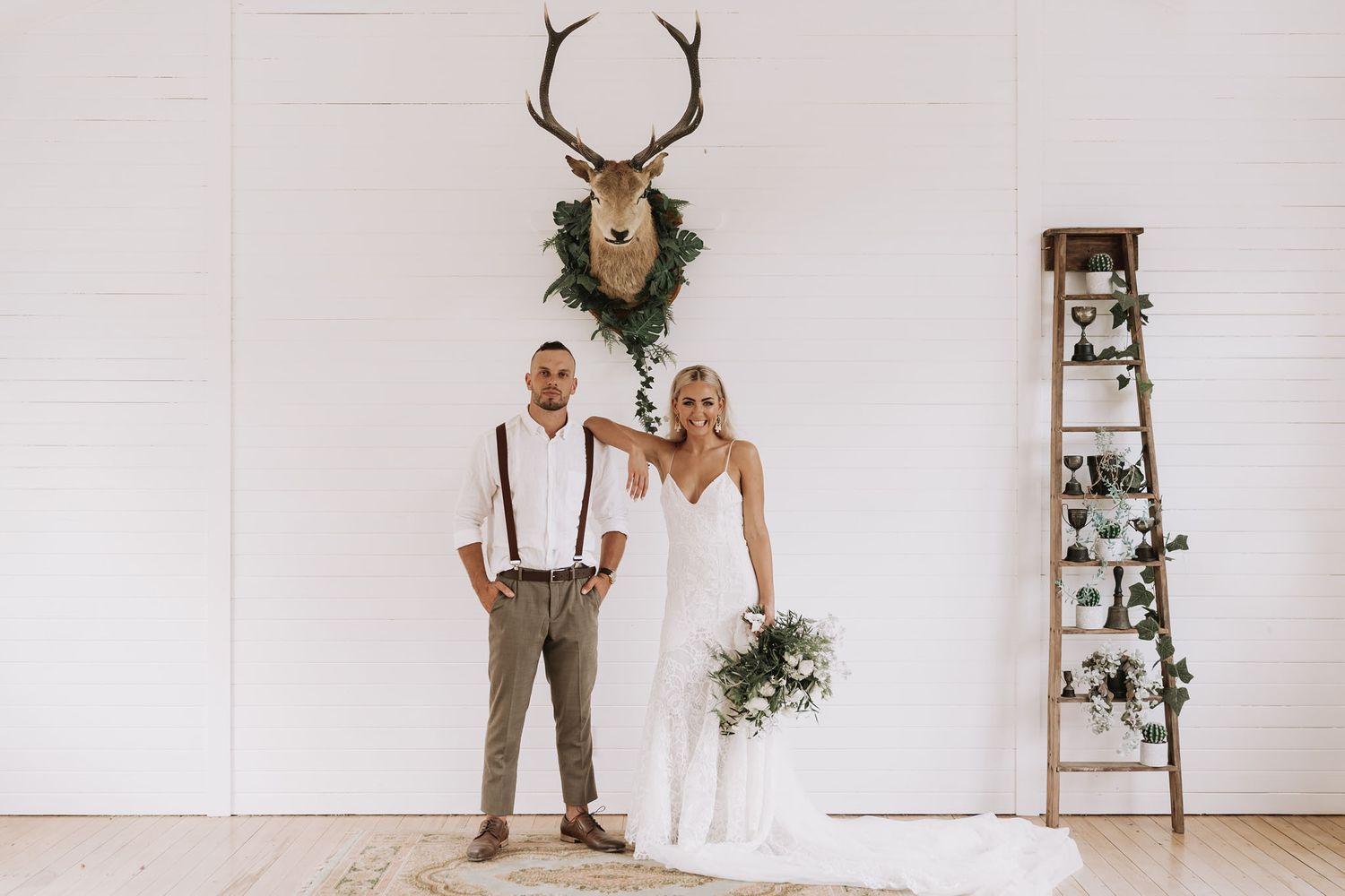 A newlywed couple stands in front of a white wall with mounted deer antlers and a rustic wooden ladder with greenery.