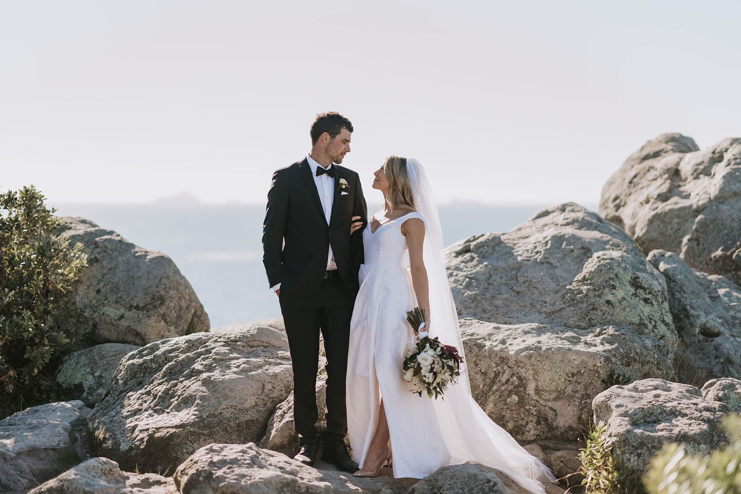 Newlyweds embrace while standing on rocky cliffs overlooking scenic mountain landscape at sunset.