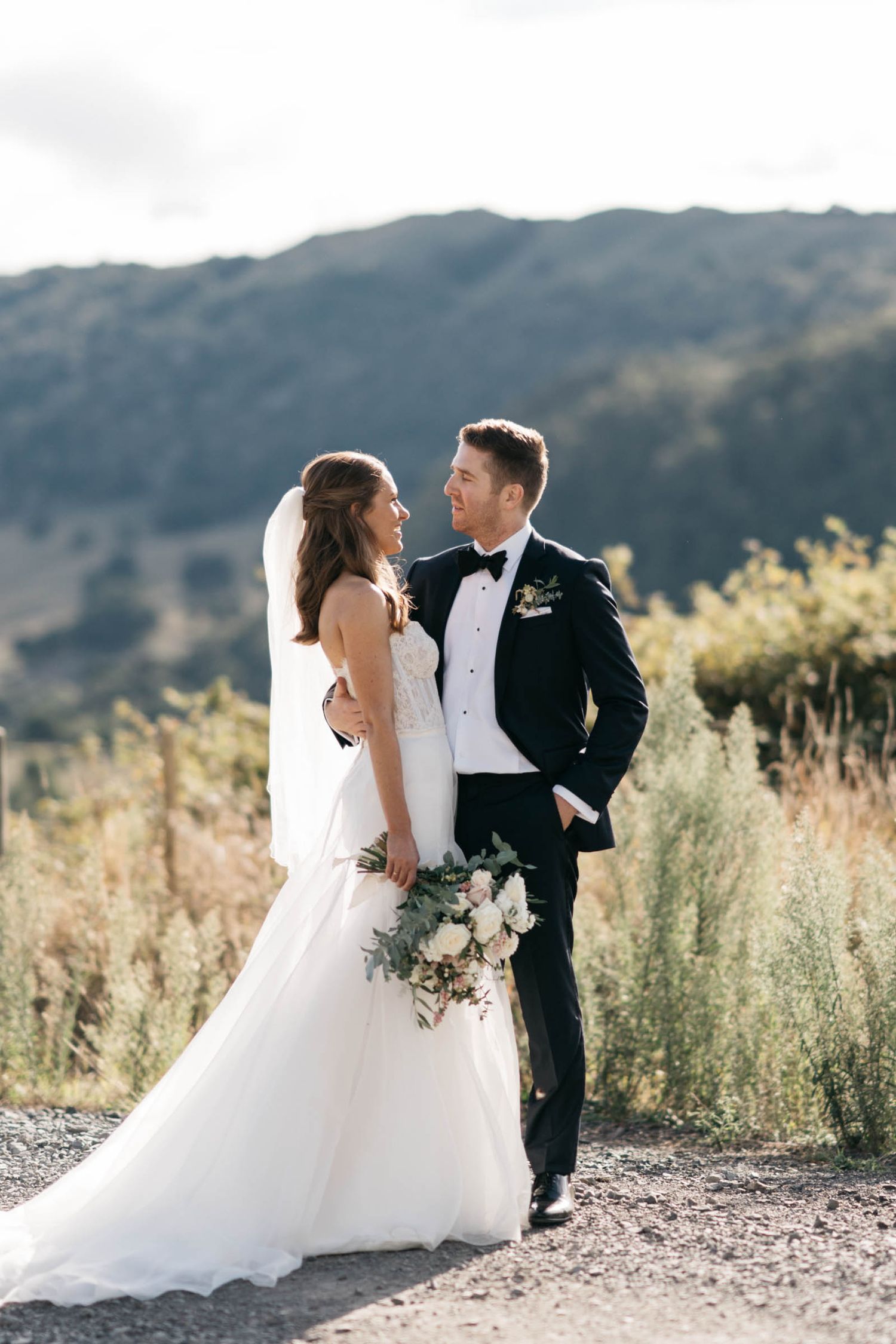 A bridal couple embraces in a romantic outdoor setting with mountains and wildflowers in the background.