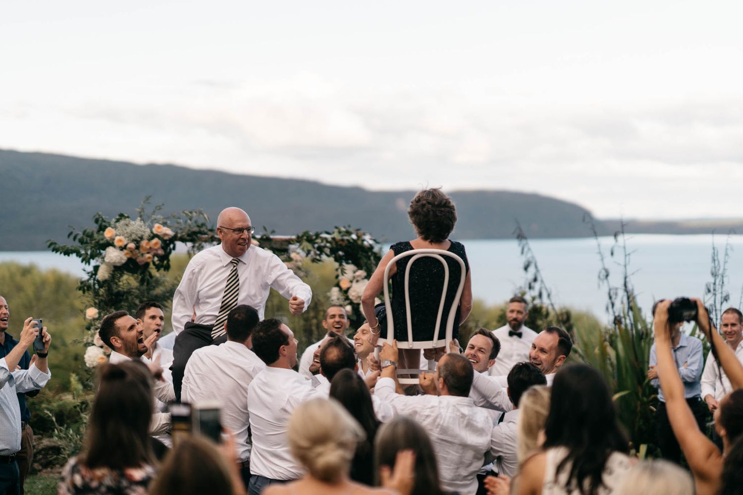 An outdoor wedding ceremony takes place on a hill overlooking a serene waterfront with mountains in the background.