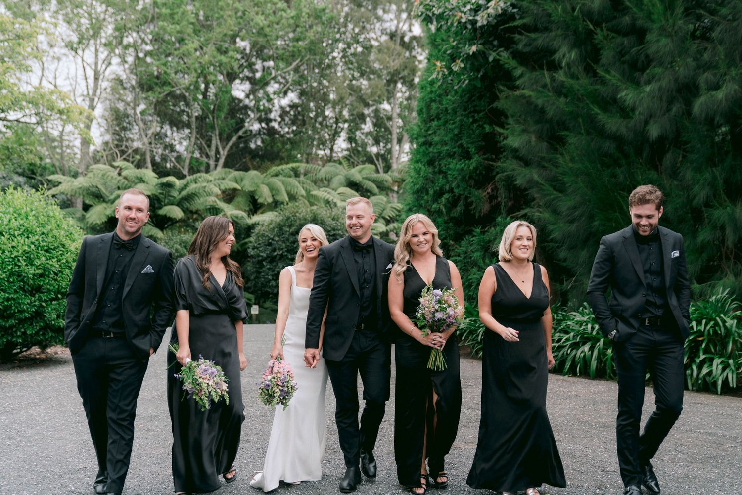Wedding party in black attire walking and laughing together on a garden path.