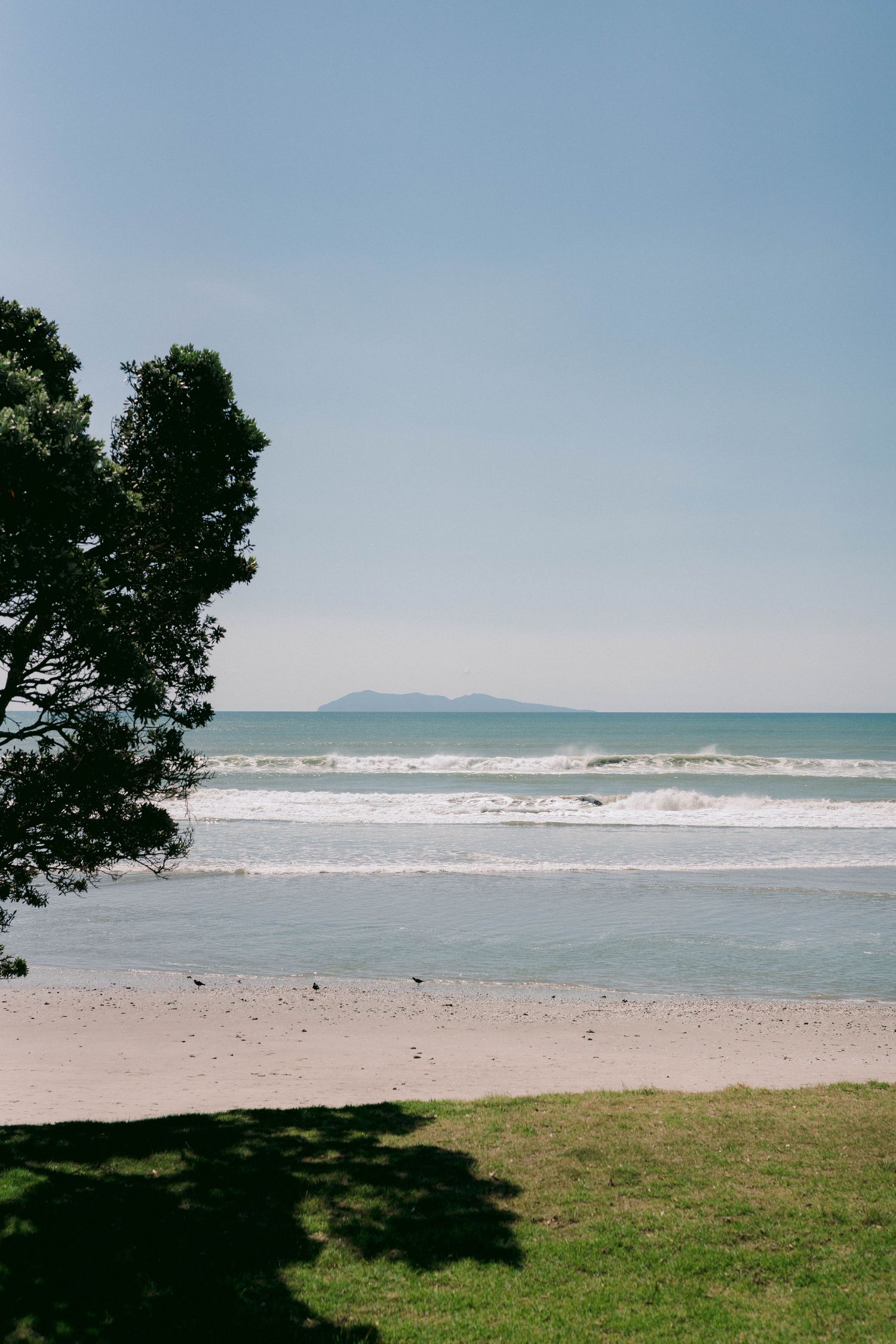Scenic beach landscape with waves crashing on shore and mountains visible in distance under blue sky.