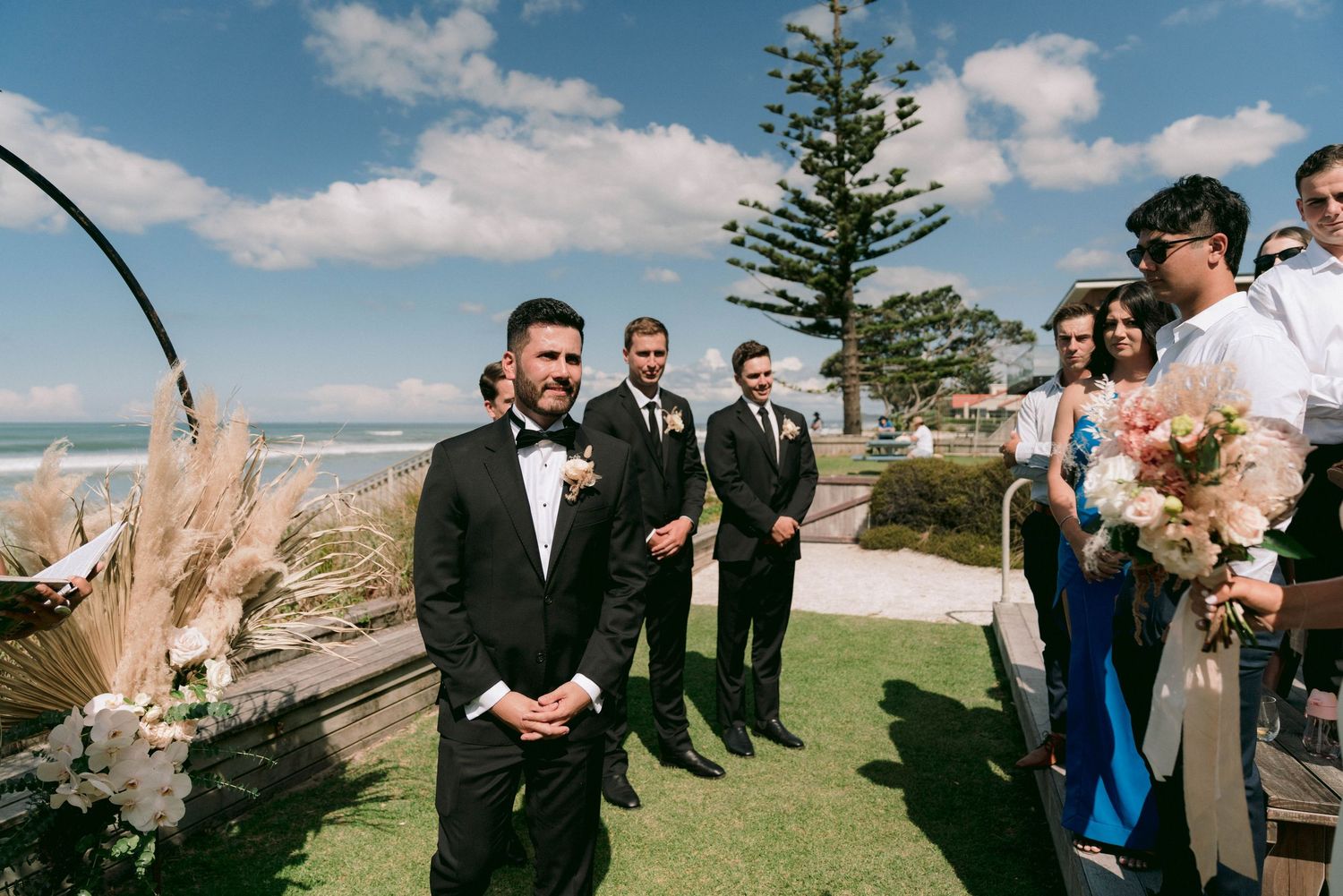 Wedding party stands on a scenic coastal cliff overlooking the ocean during a bright sunny day.