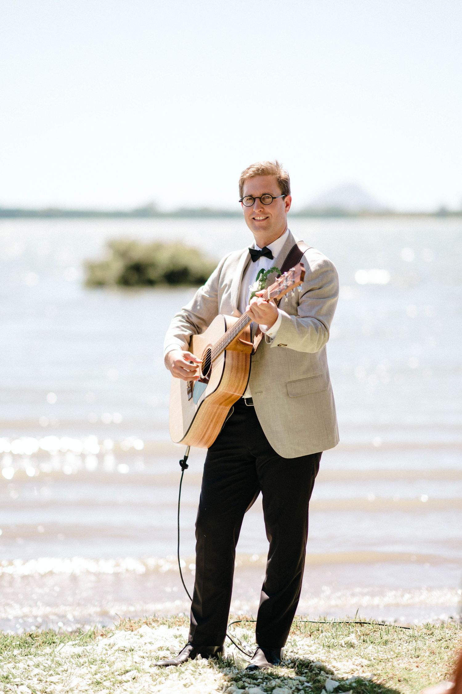 A musician in a light jacket plays acoustic guitar on a sunny beach with water and mountains in the background.