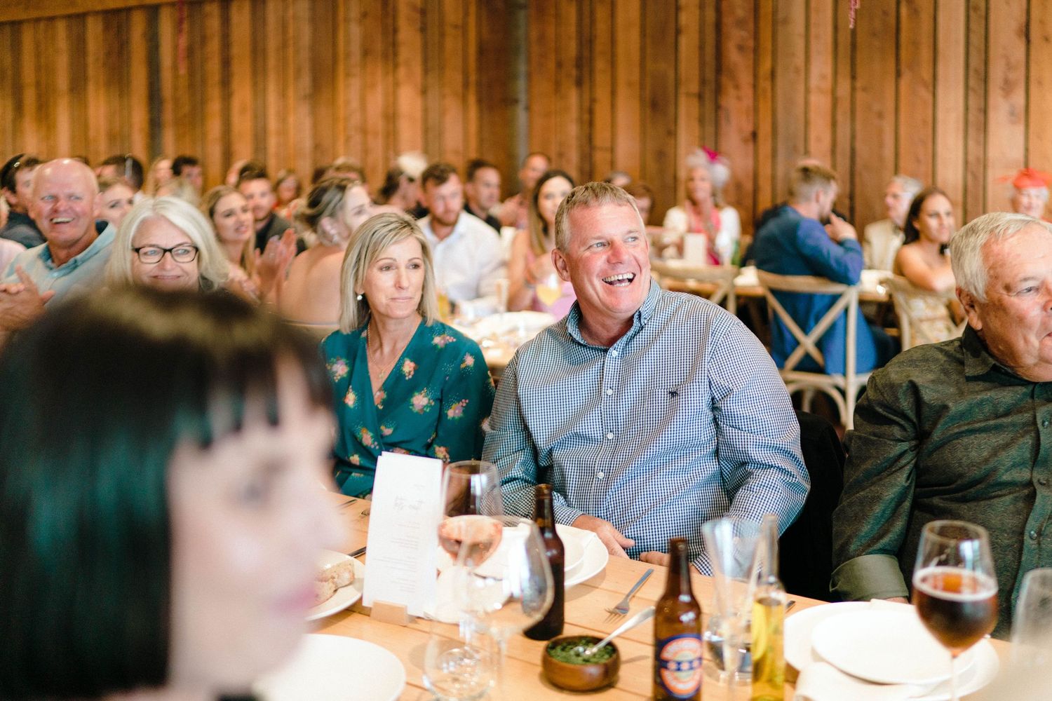Wedding reception guests seated at tables in a rustic venue with wood paneled walls.