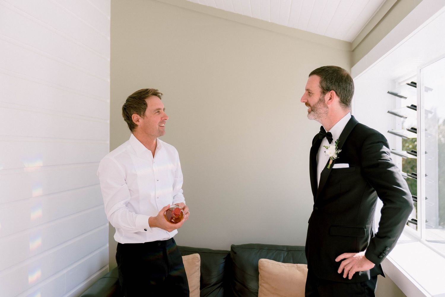 Two men in contrasting white shirt and black suit engage in animated conversation while standing in bright room.