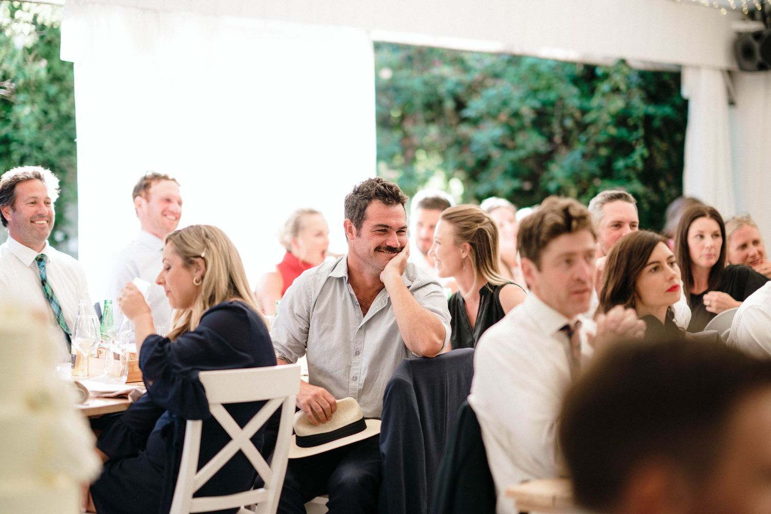 Wedding guests seated at long tables enjoying an outdoor reception dinner beneath trees.