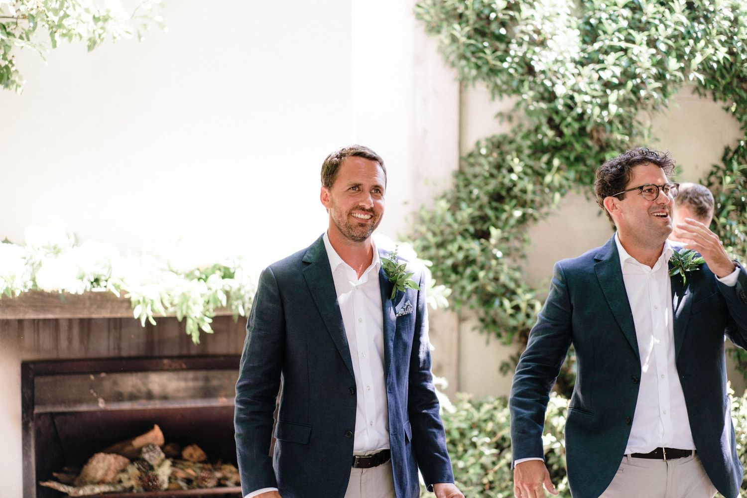 Two people wear navy blazers and white shirts while standing at an outdoor event with greenery in the background.
