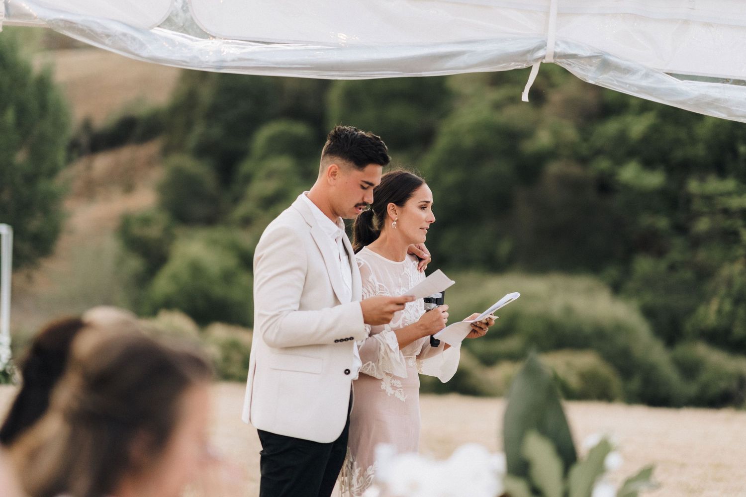 Couple reading from papers during outdoor wedding reception under white canopy.
