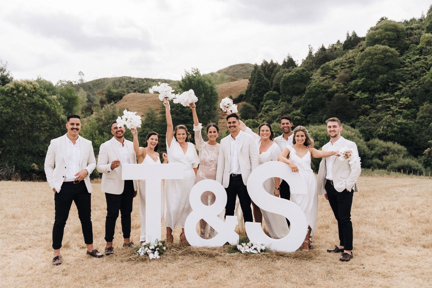 Wedding party in white outfits posing with large ampersand sign outdoors.