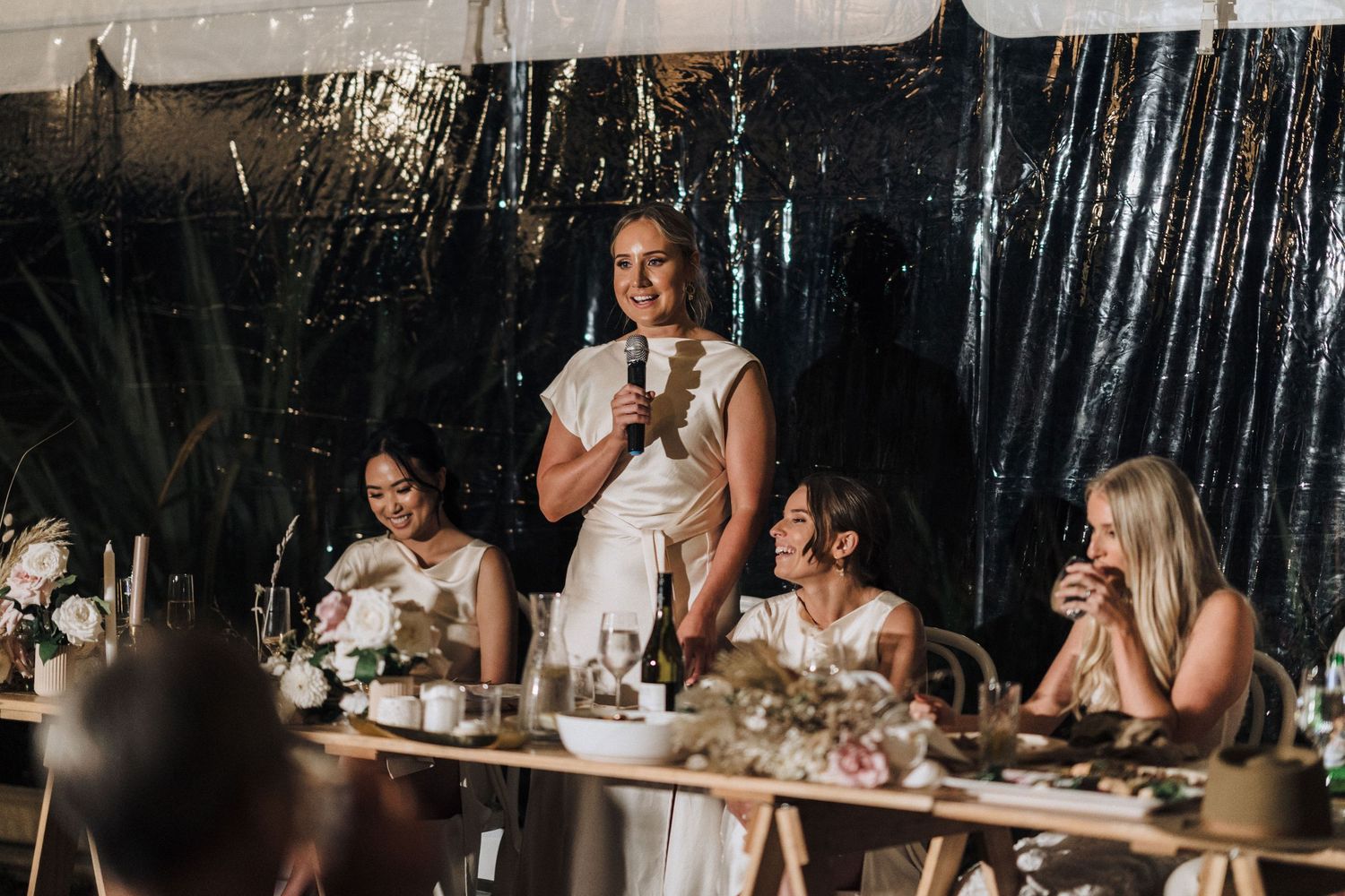 Wedding reception speech during dinner at an elegant event with sparkly black curtain backdrop and floral centerpieces.