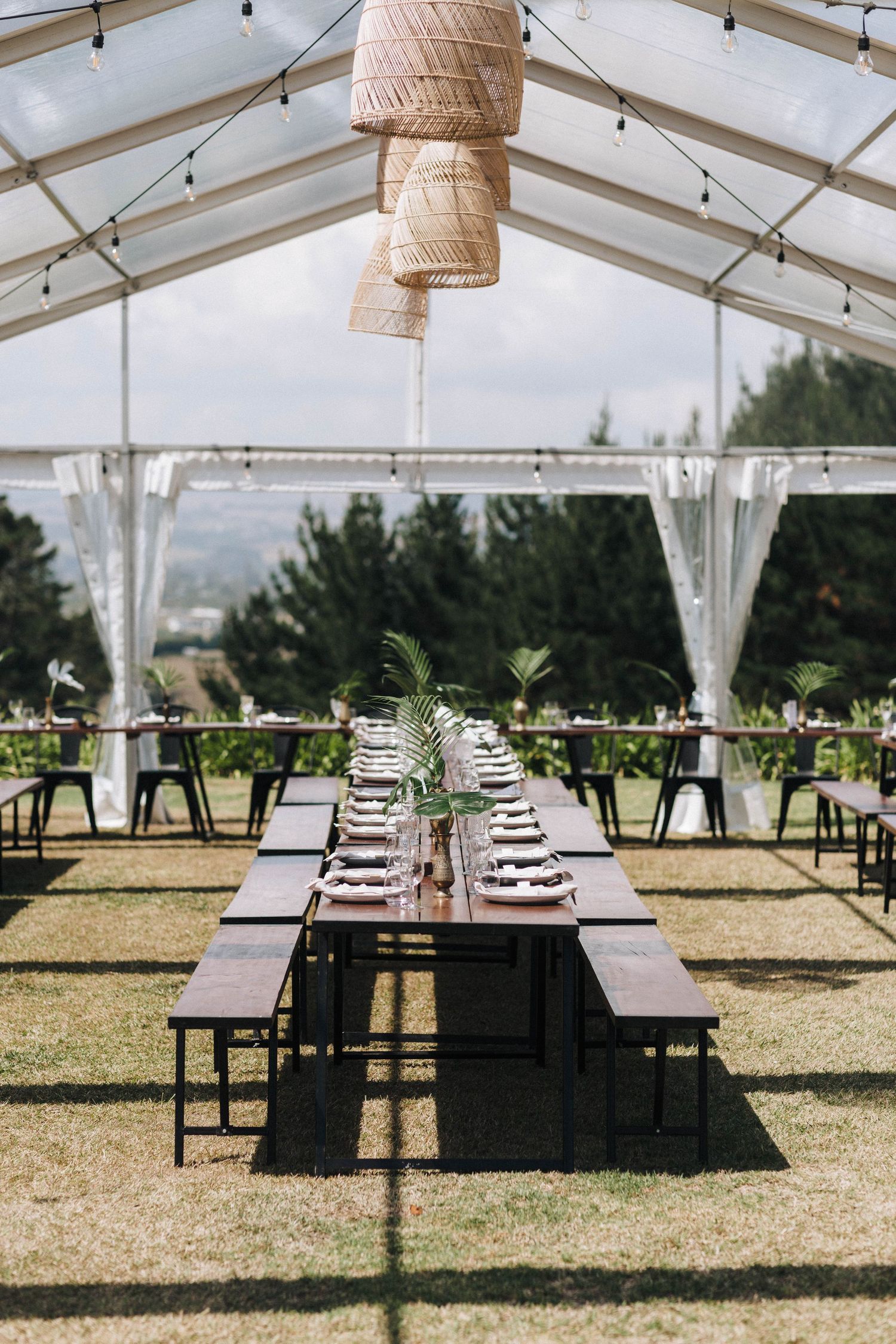 Outdoor wedding reception setup with rustic tables and woven pendant lights under a white canopy tent.