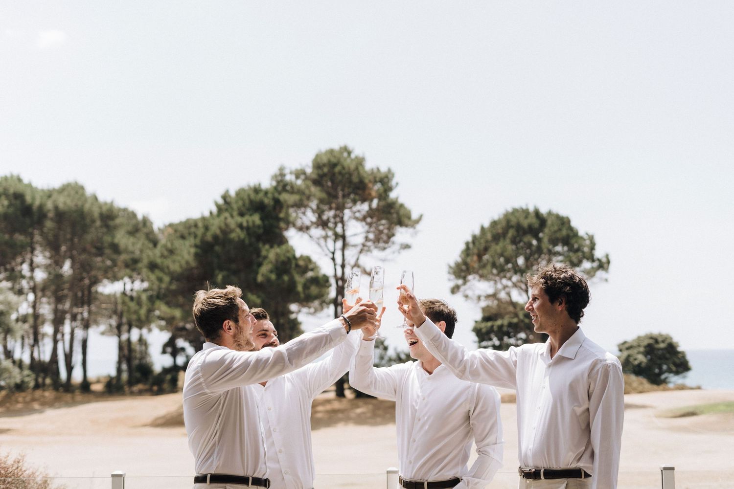 People in white attire celebrating with drinks on a sunny day with pine trees in the background.