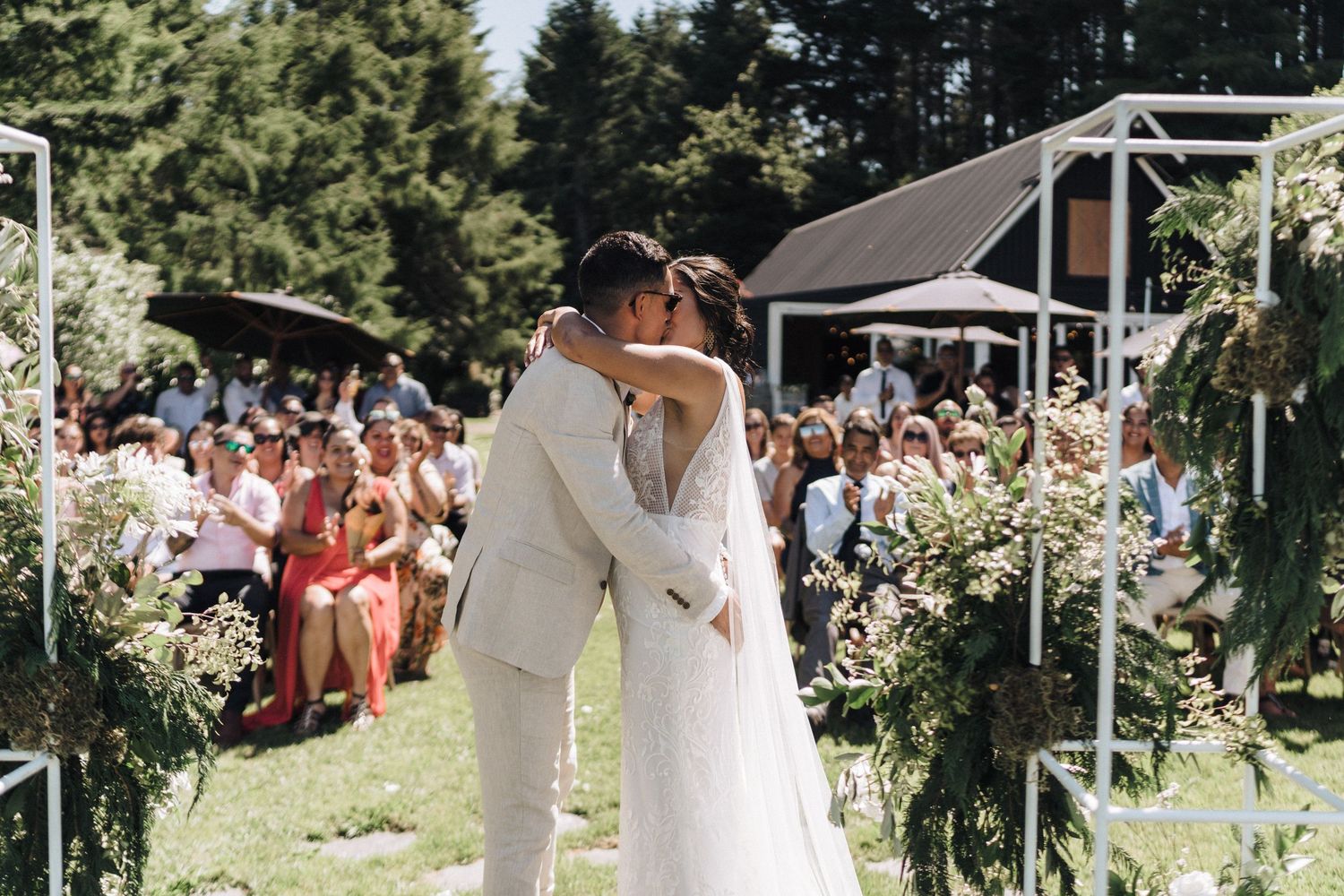 A couple shares their first kiss during an outdoor wedding ceremony surrounded by guests and floral decorations.