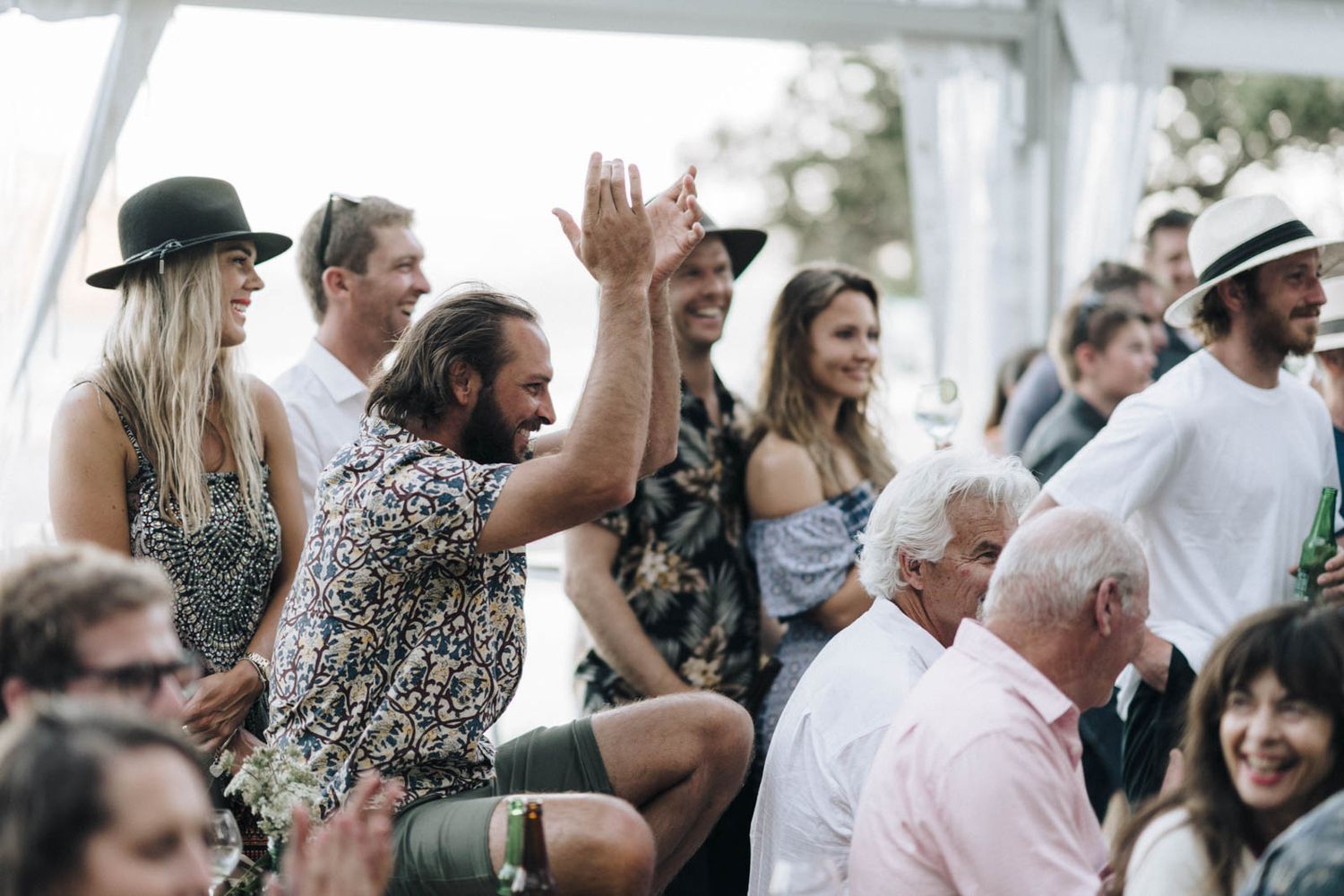 Wedding guests enjoying an outdoor celebration under a white tent with some standing and raising hands in excitement.