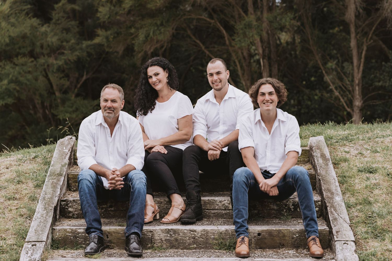 Four individuals sit casually on rustic outdoor wooden steps against a wooded background.