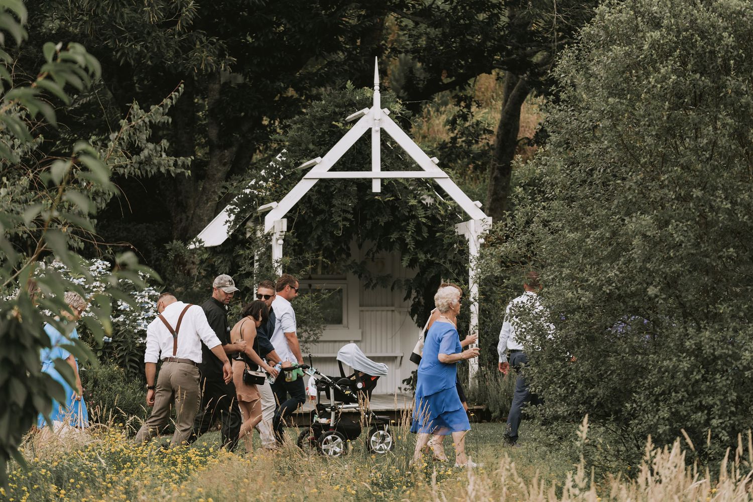 A rustic outdoor wedding ceremony setup with a white wooden arch in a garden surrounded by trees and greenery.