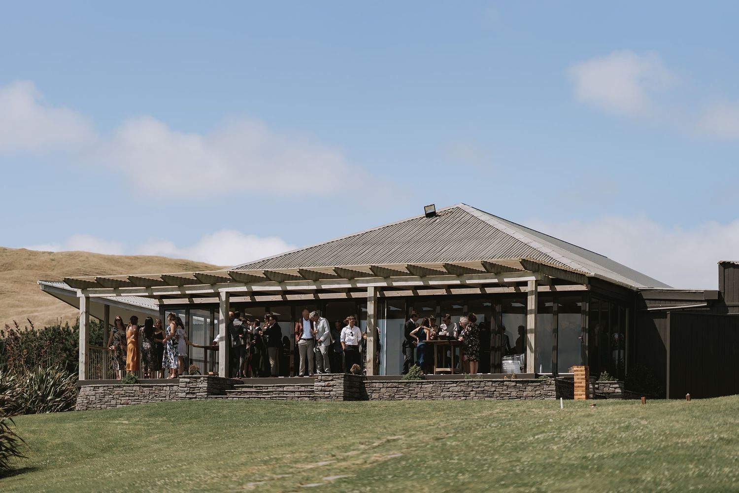 A rustic outdoor pavilion venue with a gray roof stands against rolling hills on a sunny day.