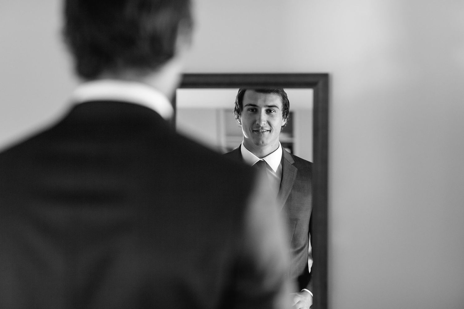 Black and white photo showing reflection in mirror of someone adjusting their suit and tie.