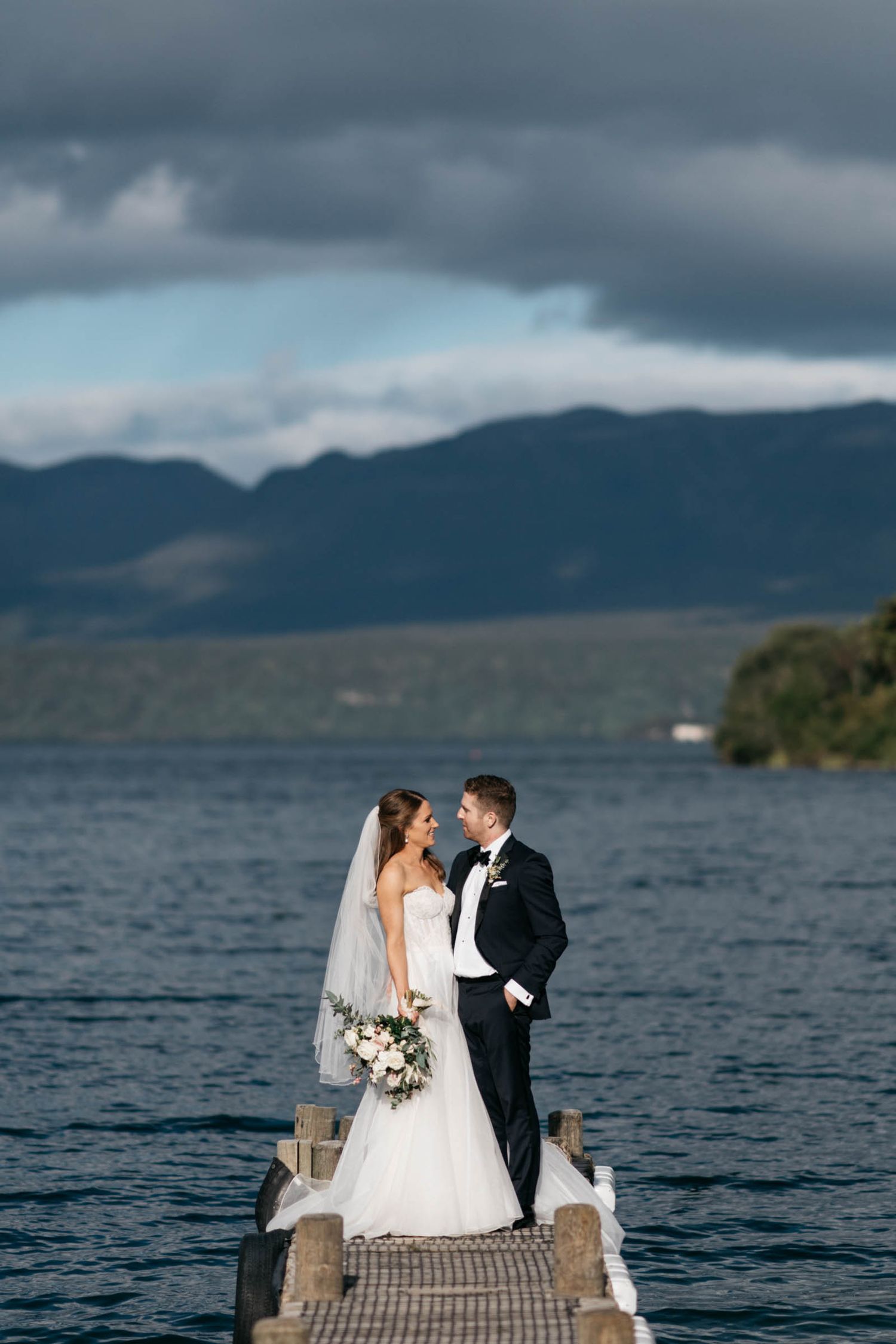 A couple standing together on a wooden dock overlooking a scenic lake with mountains in the background during a cloudy day.