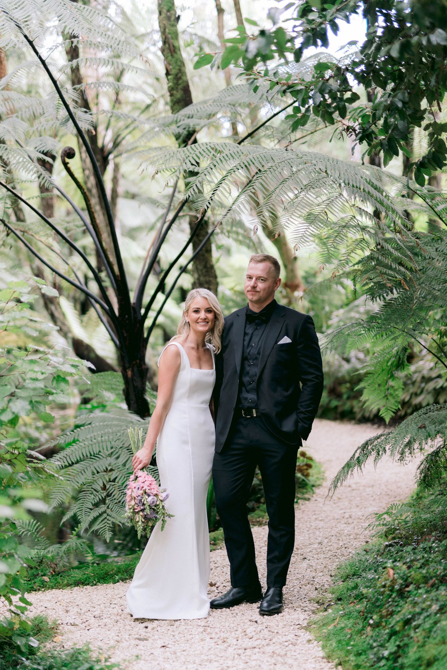 A couple poses on a garden path surrounded by ferns and greenery at their wedding.
