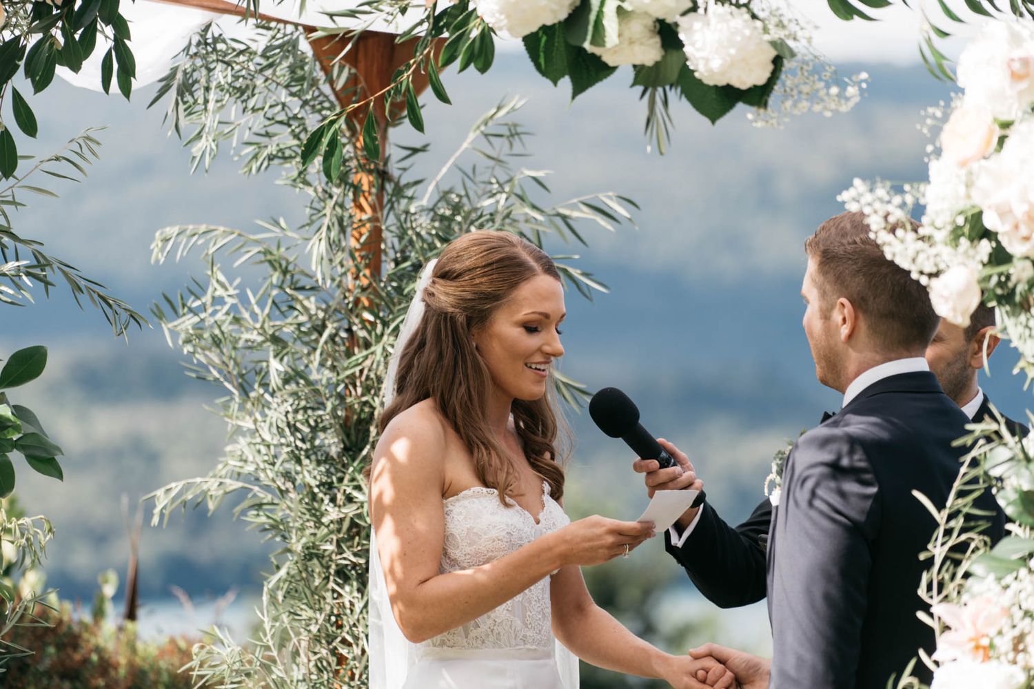 Under a floral arch at an outdoor wedding ceremony, a couple exchanges heartfelt vows surrounded by nature.