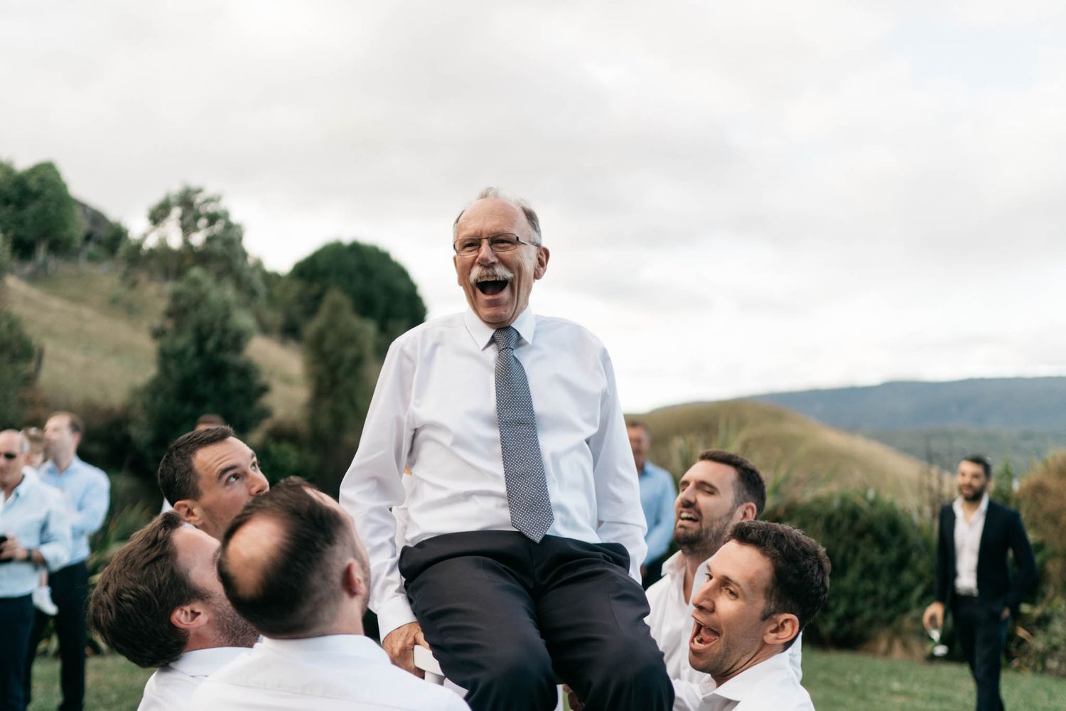 A person in white dress shirt and tie lifted up in celebration by a group of people at an outdoor wedding party.