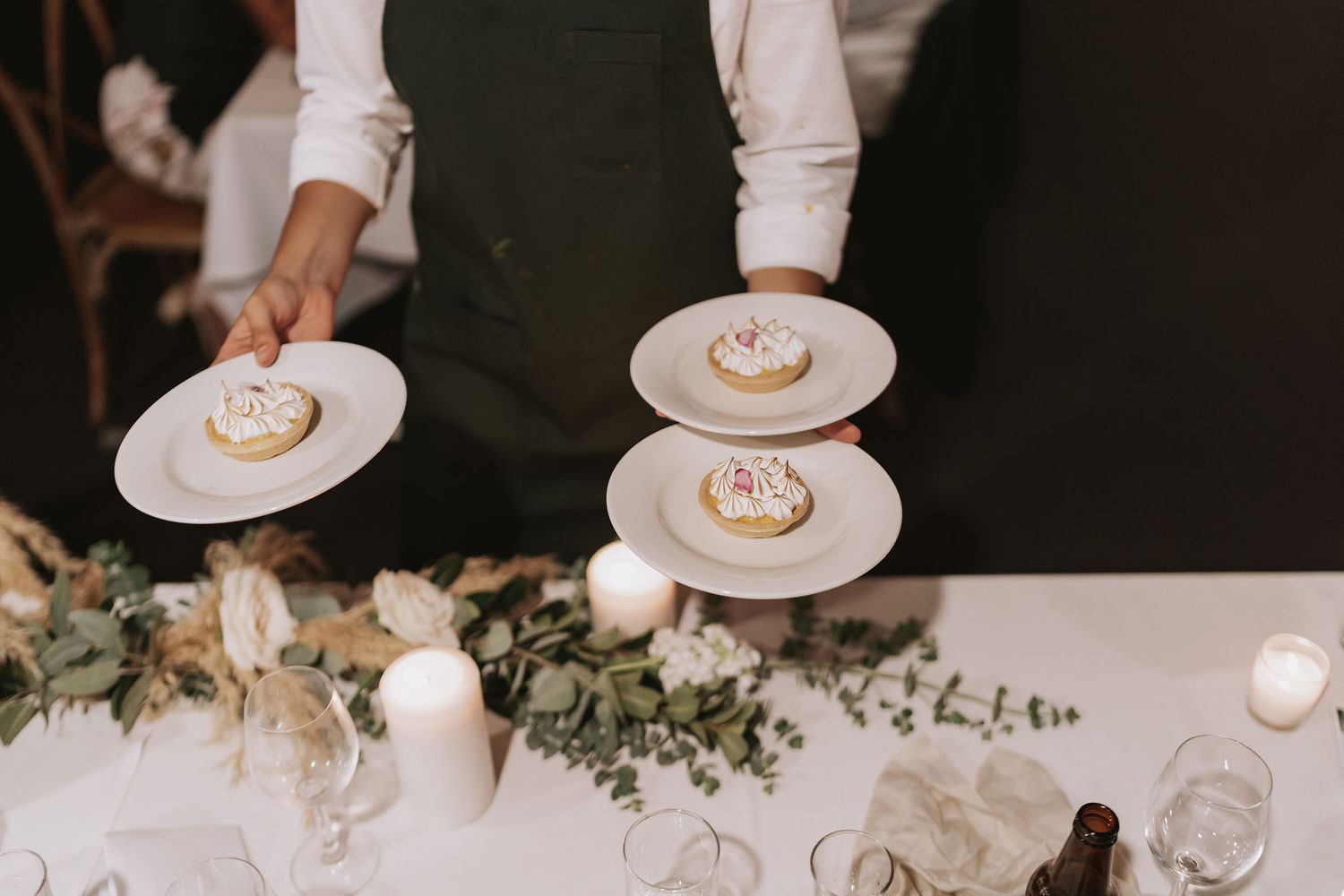 Waiter serves elegant dessert plates at a wedding reception table decorated with eucalyptus and white candles.