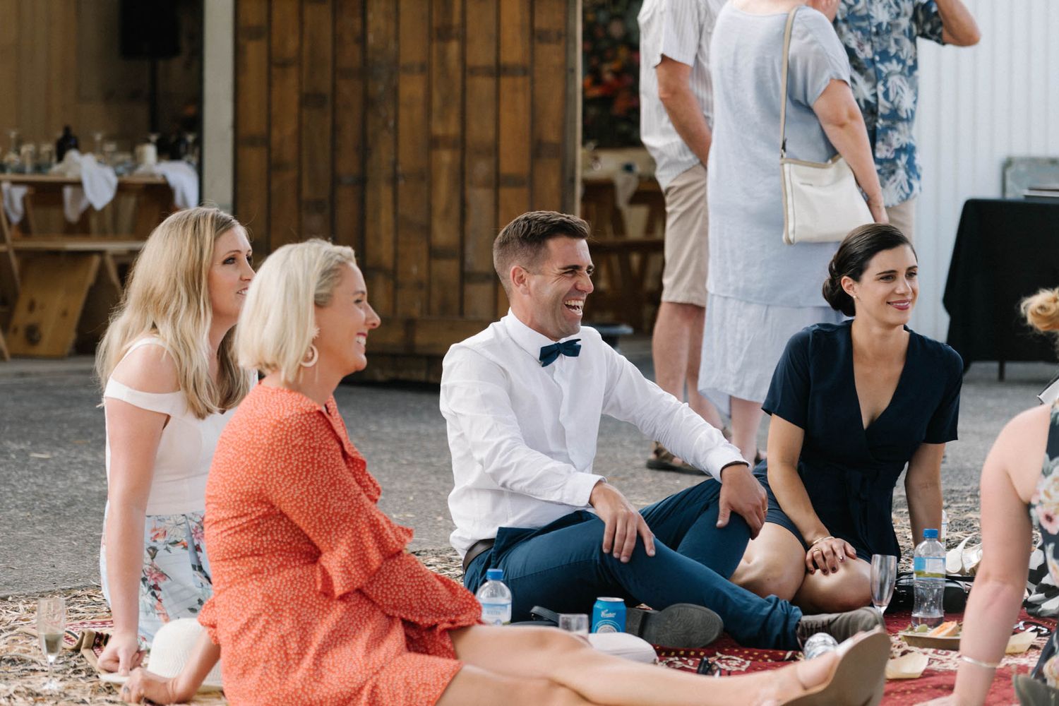 Group of people sitting casually on a blanket at an outdoor gathering with rustic wooden doors in background.