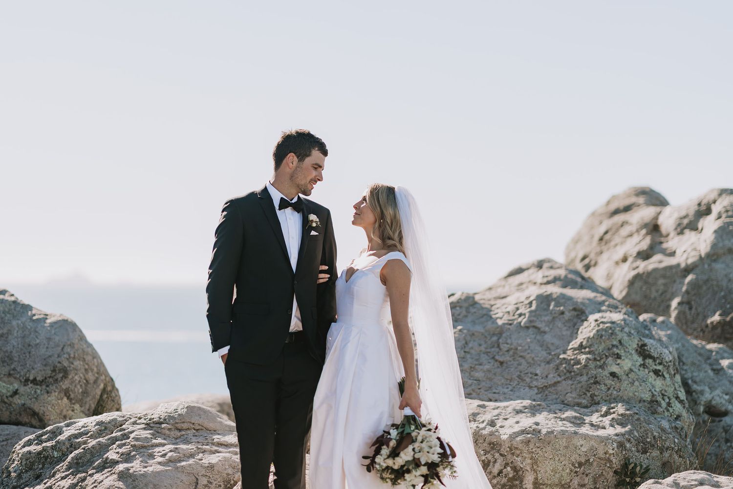 A couple in formal wedding attire stands together on a rocky coastal cliff overlooking an ocean at sunset.