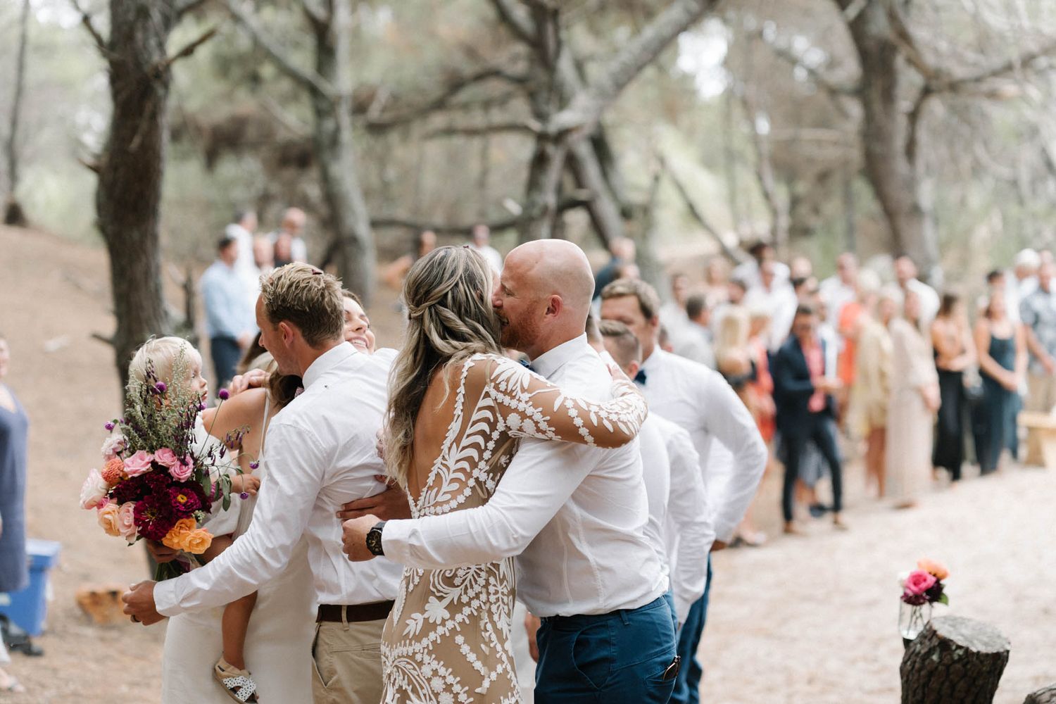 Wedding guests dance together on a wooded path during an outdoor reception celebration.