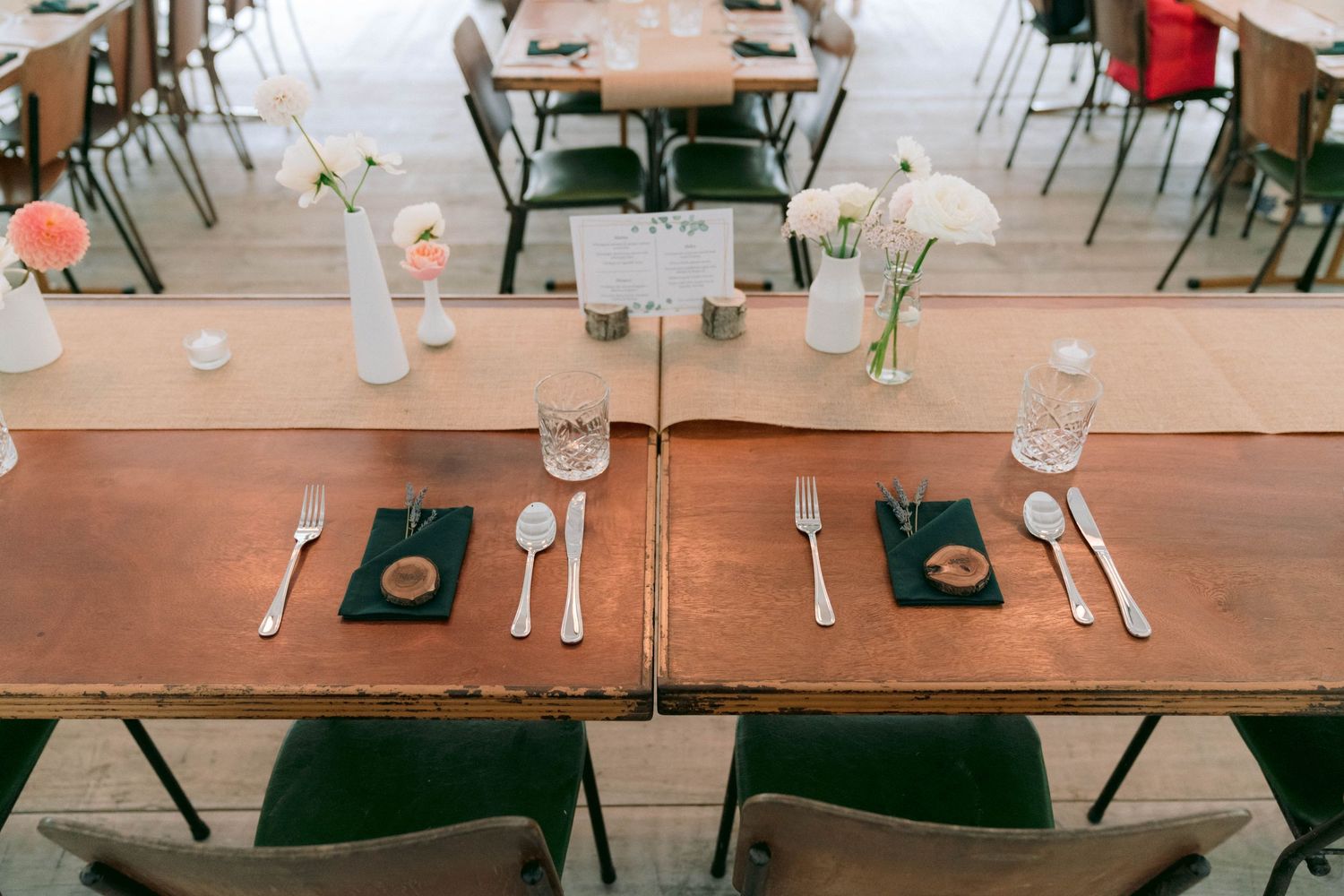 Simple place settings with silverware and green coasters on wooden tables with green chairs in a dining hall.