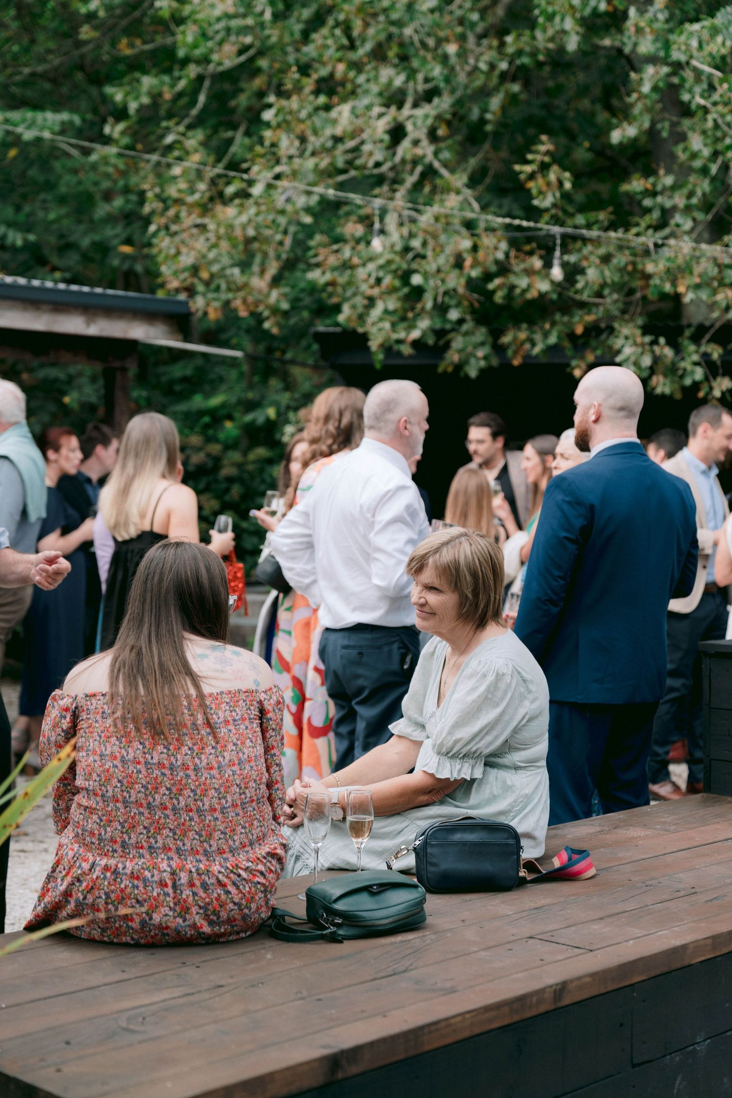 Wedding guests relax and socialize on wooden benches during an outdoor reception.
