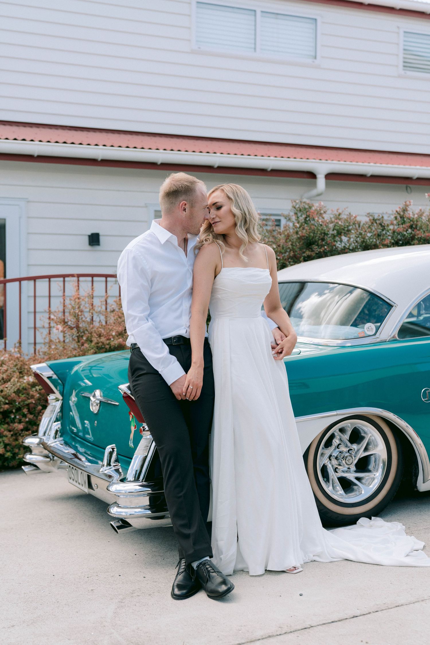 A wedding couple poses beside a vintage turquoise car in front of a white house.