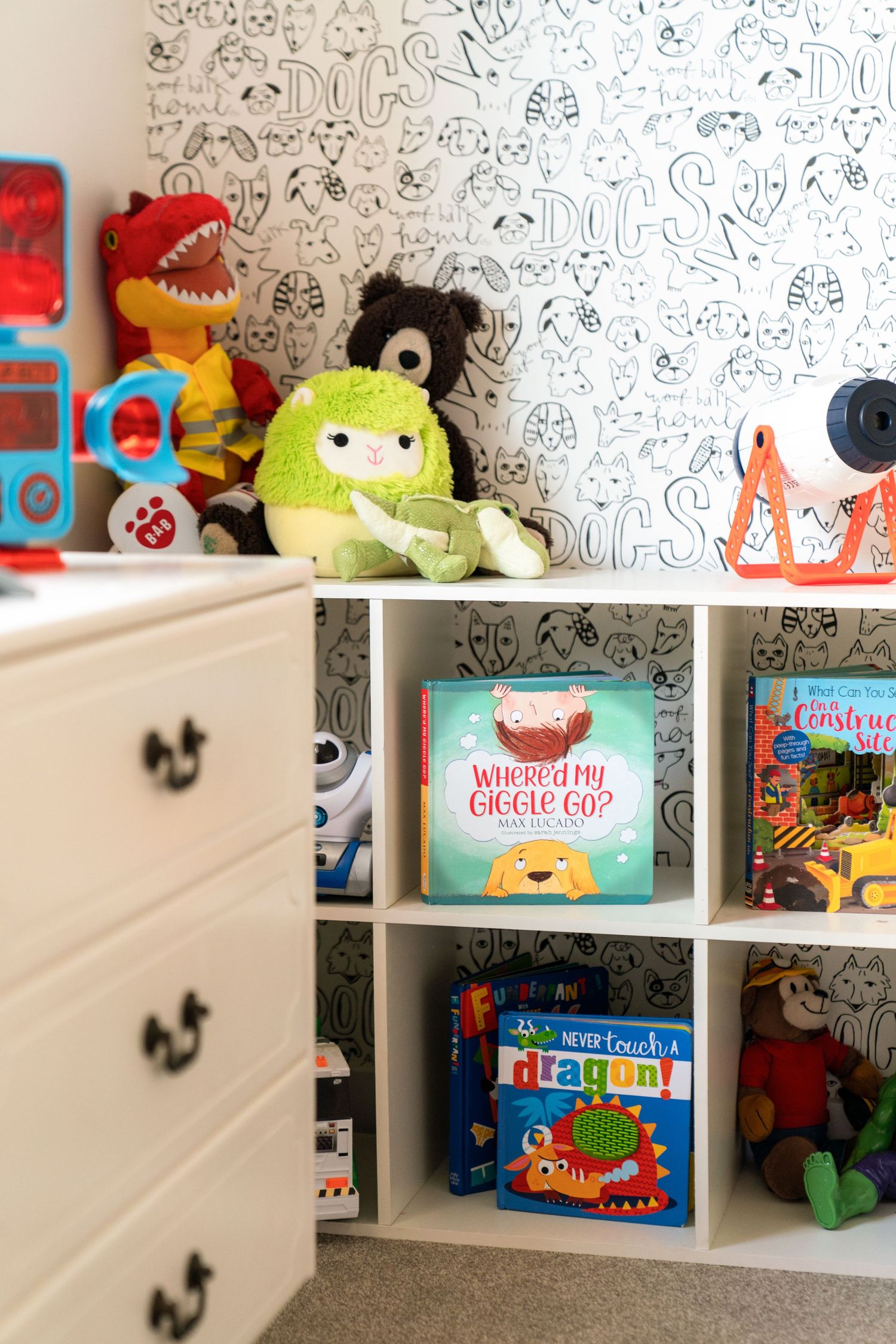 Children's bedroom shelf featuring colorful stuffed animals and books against a patterned wallpaper with white drawers.