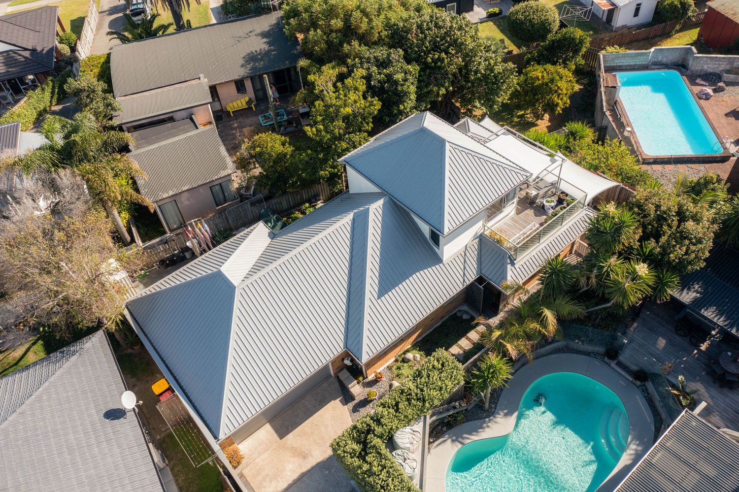 Aerial drone view of modern house with grey roof and swimming pool in backyard surrounded by trees.