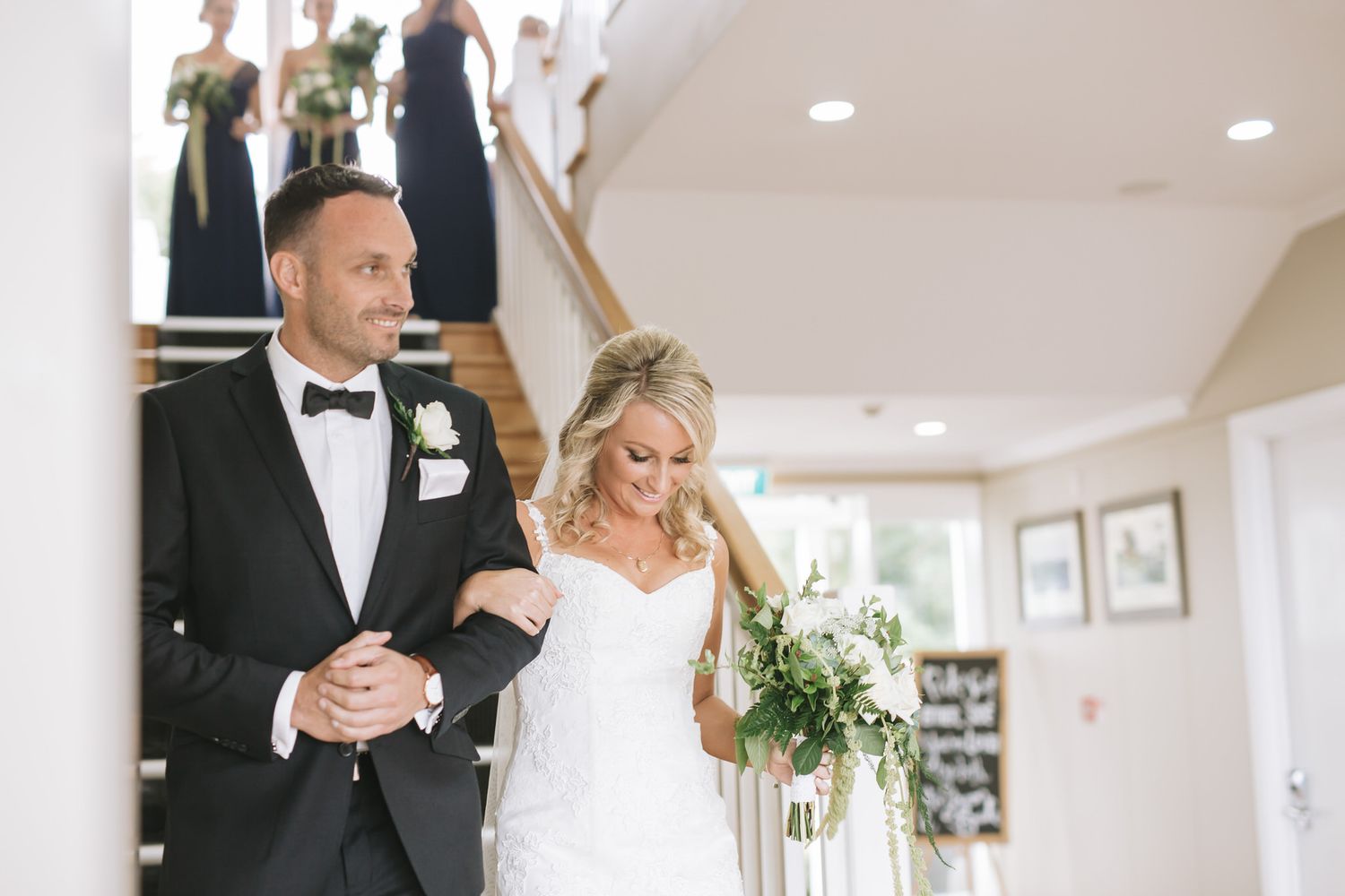 Newly married couple walks joyfully down hallway with bride in white dress holding bouquet and groom in tuxedo.