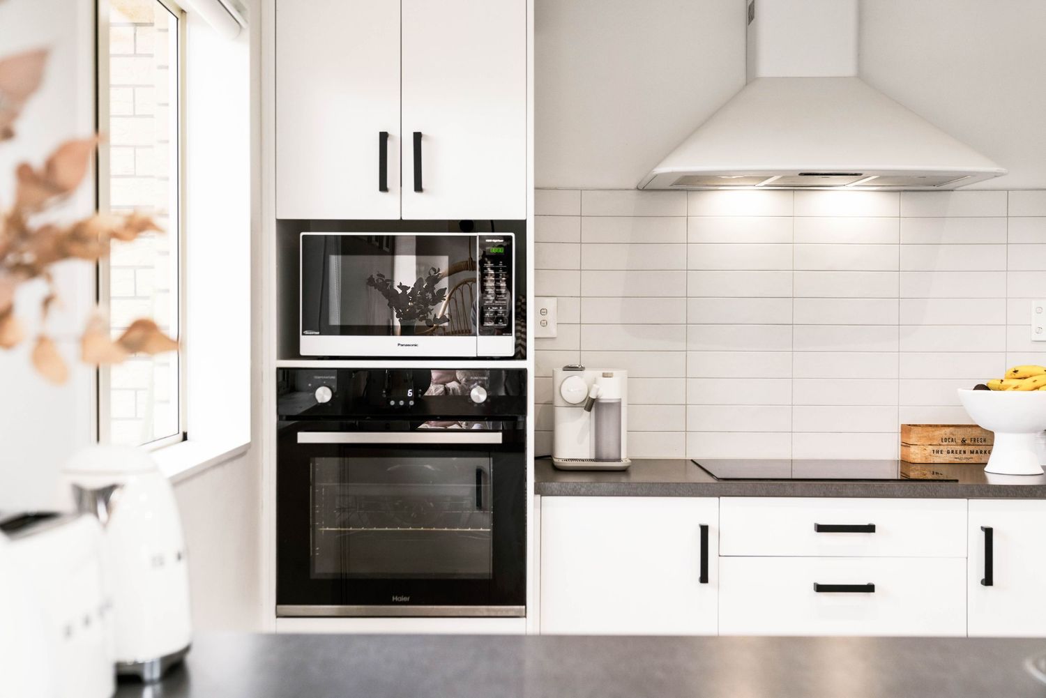 Modern white kitchen with built-in microwave, oven, and range hood featuring white subway tile backsplash and dark countertops.
