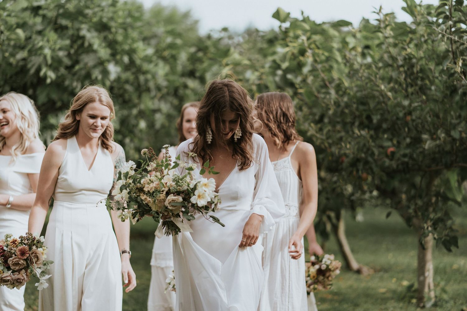 A group of bridesmaids in flowing white dresses walk together holding floral bouquets in an orchard garden setting.