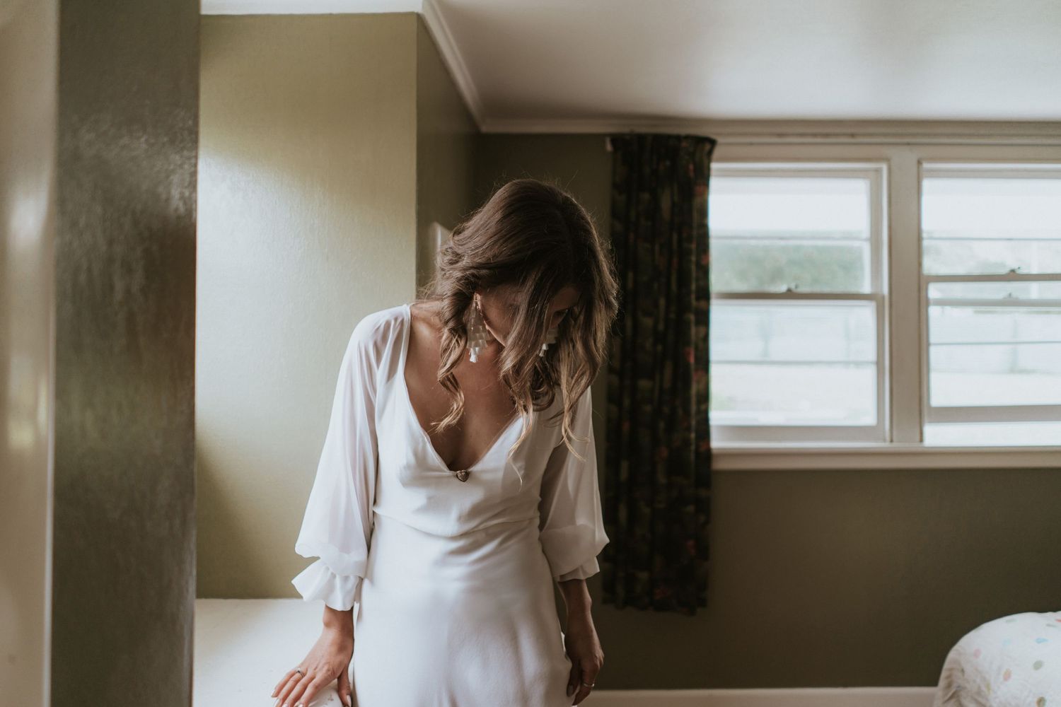 Person in a flowing white dress with long sleeves stands by a window in a minimalist room interior.