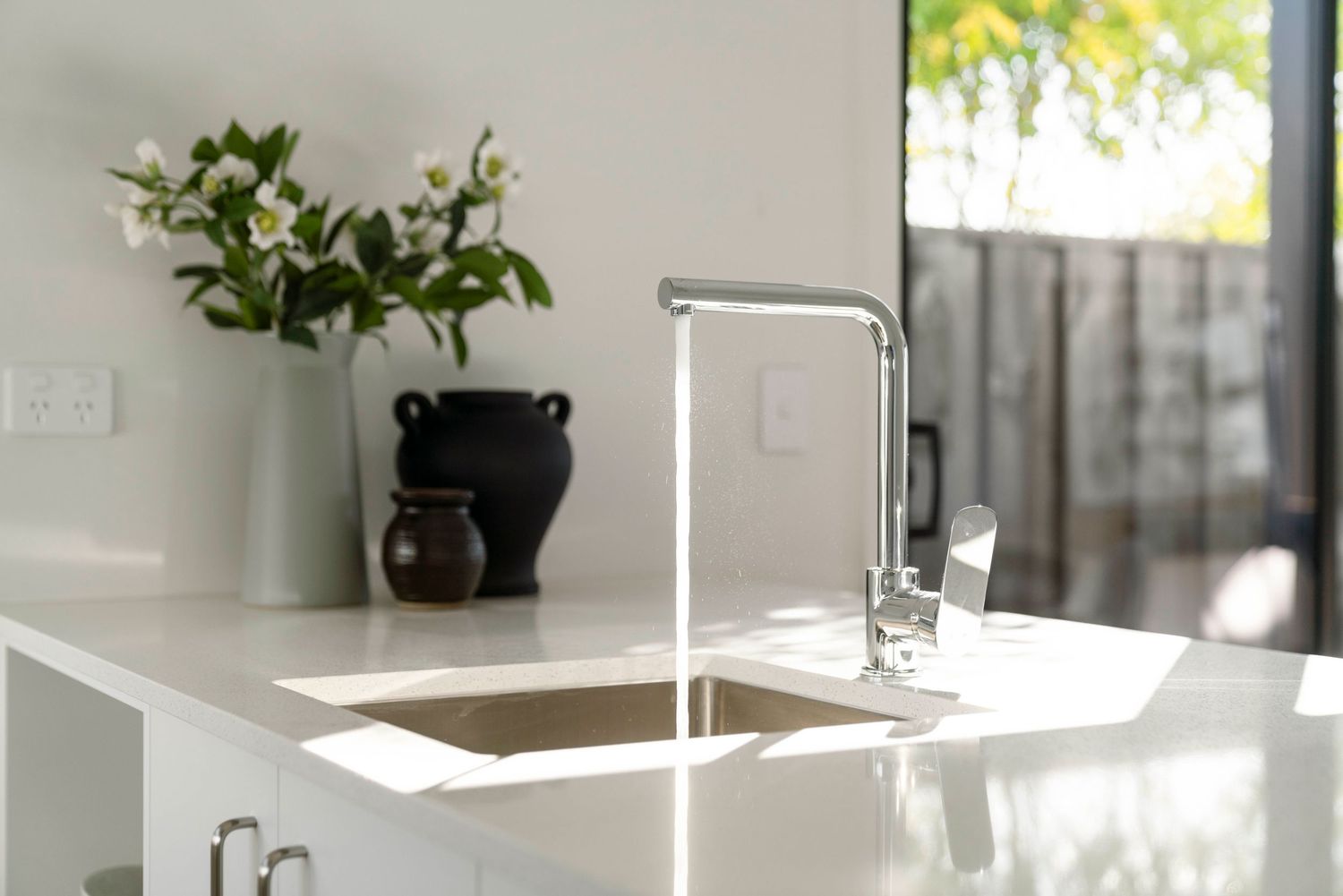 Modern kitchen with sleek white countertop featuring a minimalist chrome faucet and decorative plants in white vase.