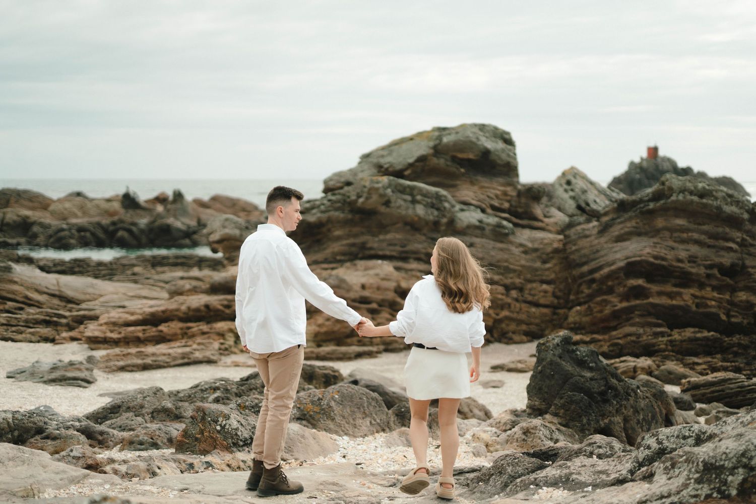 Two people wearing white hold hands while standing on rocky coastal terrain during an outdoor photoshoot.